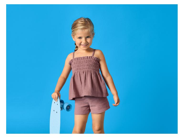 Usmívající se dívka v topu a šortkách barvy slézu, držící světle modrý skateboard.