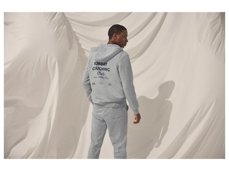 Muž v šedé mikině s kapucí a teplácích s textem „SUNDAY COUCHING Club“ na zádech.