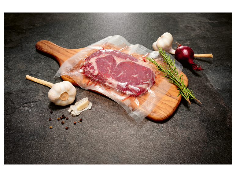 Vakuově balený hovězí steak na dřevěné prkénku s česnekem, cibulí a rozmarýnem.