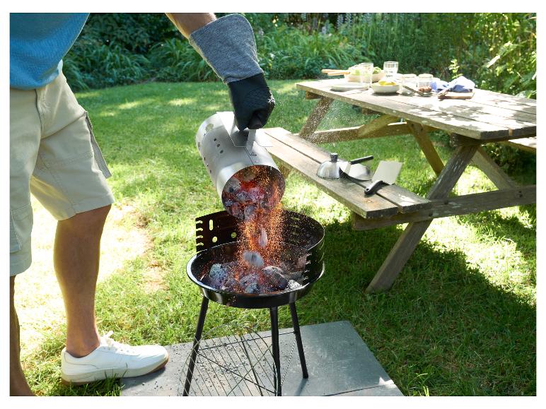 Muž sype žhavé uhlí z podpalovacího komínu do grilu, vedle piknikový stůl s nádobím.