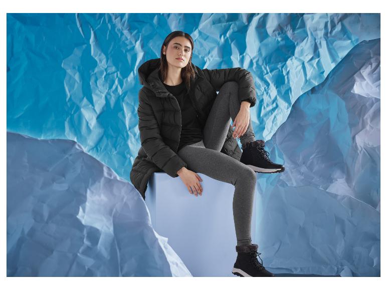 Žena v černé bundě a šedých legínách, sedící na modré kostce, na modrém pozadí.