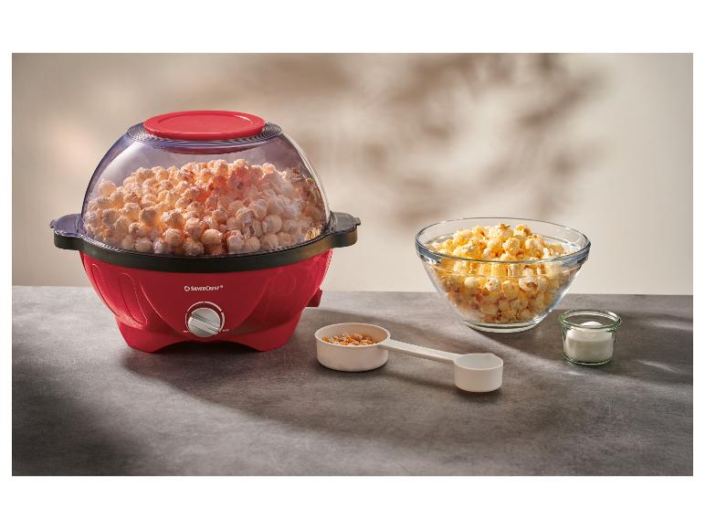 Červený popcornovač plný popcornu, s miskou popcornu a přísadami na stole.