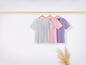 Tři trička v šedé, růžové a fialové barvě visící na dřevěné ramínku.