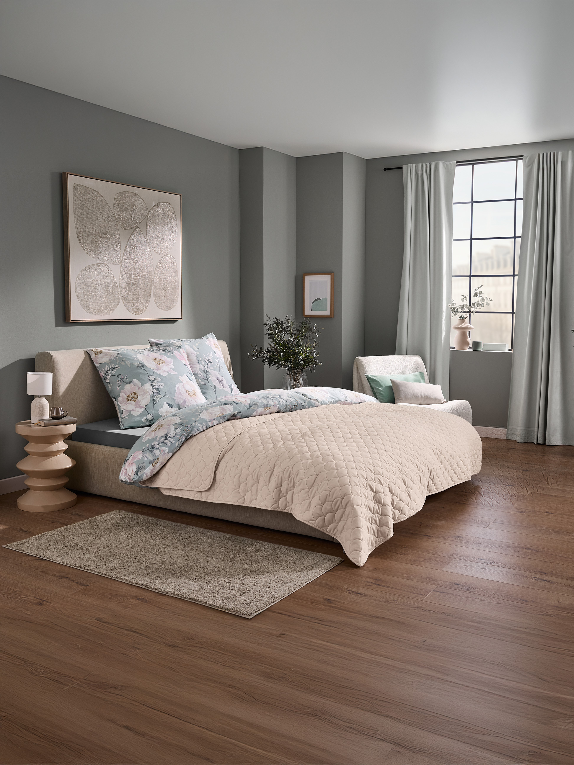 Ložnice s postelí s květinovým povlečením a béžovým prošívaným přehozem a oknem se záclonami.