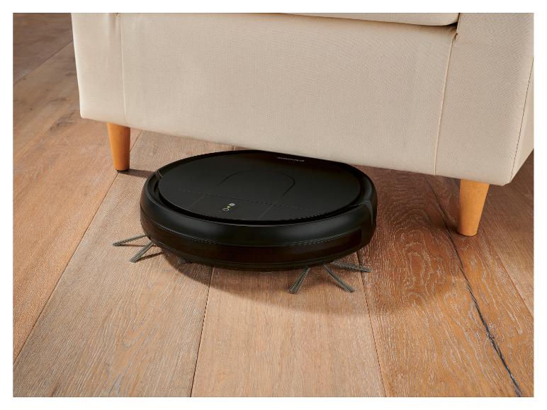 Černý robotický vysavač pod béžovou pohovkou na dřevěné podlaze.