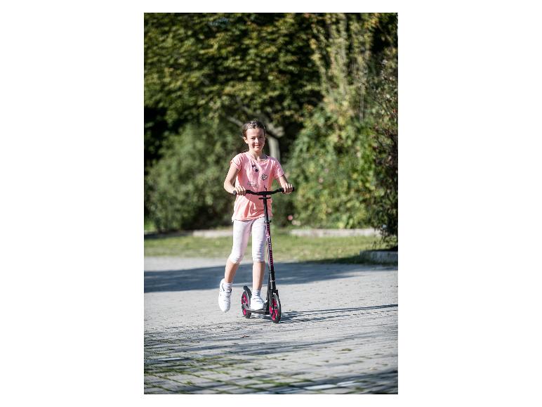 Usmívající se dívka v růžovém tričku a bílých capri kalhotách, jedoucí na černo-růžové koloběžce.