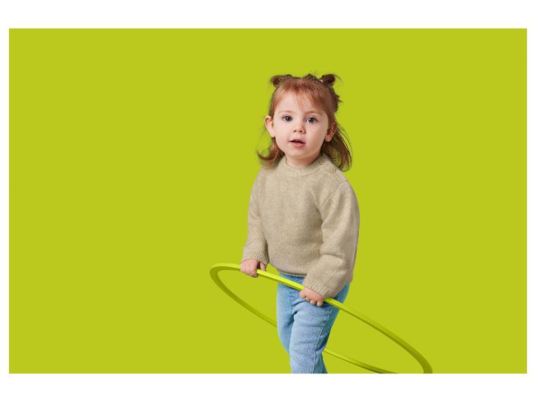 Batole s rusými vlasy, v béžovém svetru a džínách, drží zelenou obruč.