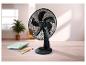 Černý stolní ventilátor na dřevěném stole s poznámkovými bloky a pery, ve světlé místnosti.