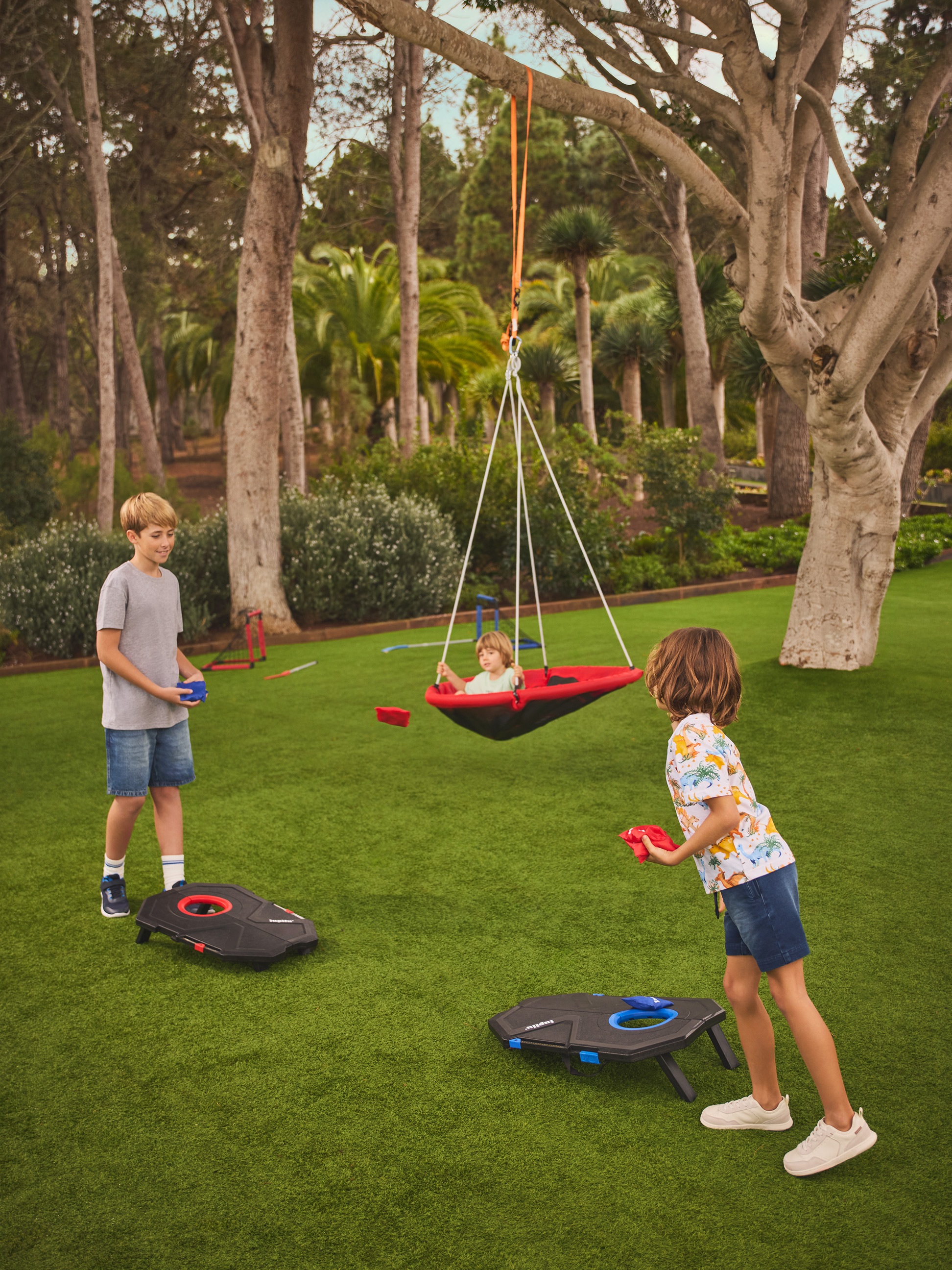 Dvě děti hrají cornhole na trávníku, zatímco třetí se houpe v hnízdní houpačce.