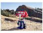 Hračka Optimus Prime stojí na skalnatém, písčitém terénu pod jasnou oblohou.