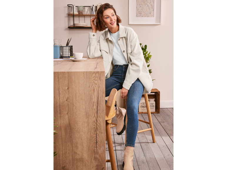 Žena v světle modrém tričku, bílé bundě a modrých úzkých džínách.