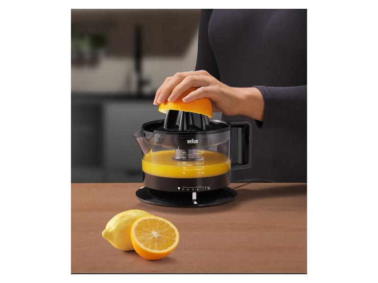 Žena odšťavňuje pomeranč citrusovačem, s citrony a pomeranči na dřevěném stole.