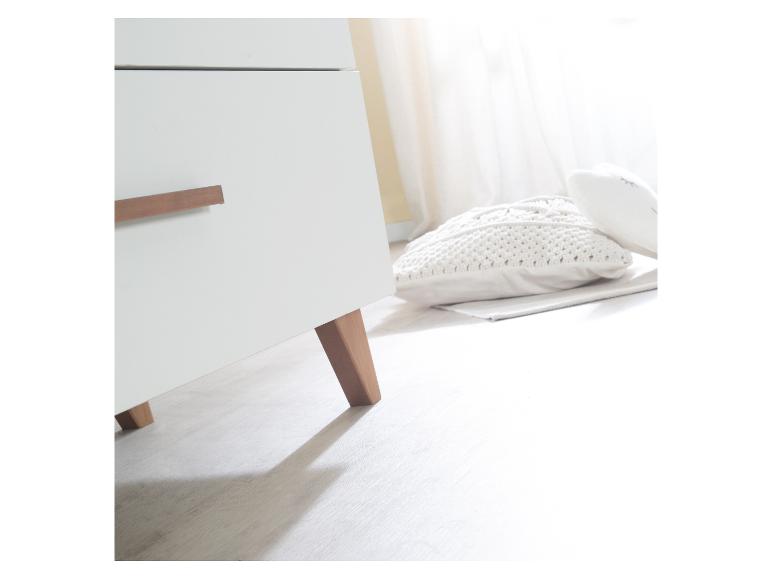Bílá komoda s dřevěnými úchyty a nohami, vedle pleteného polštáře na podlaze.
