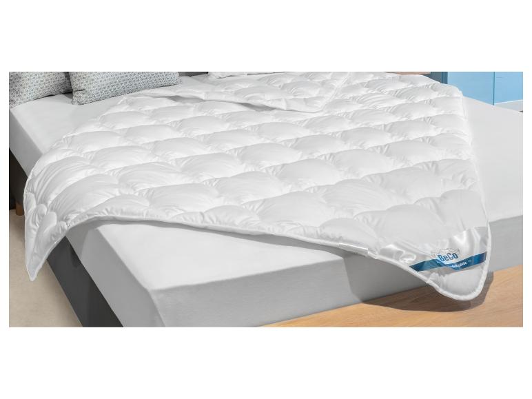 Bílá prošívaná přikrývka BeCo na posteli se vzorovanými polštáři.