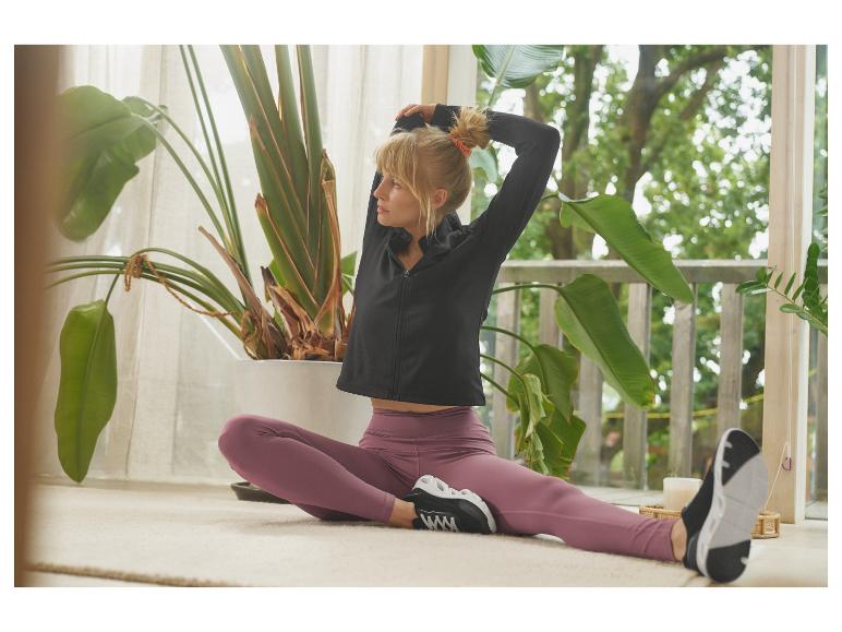 Žena v černé sportovní bundě a fialových legínách se protahuje uvnitř.