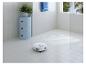 Robotický vysavač uklízí dlážděnou koupelnu s modrou skříňkou a rostlinou.