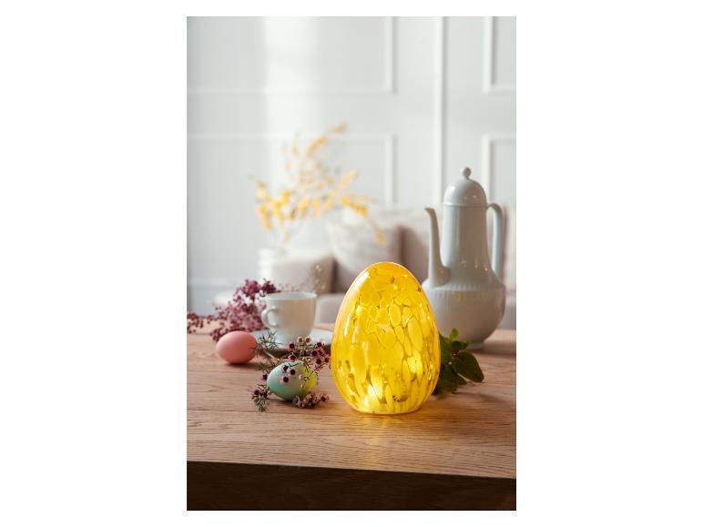 Žlutá skleněná lampa ve tvaru vajíčka na dřevěném stole s vejci a květinami.