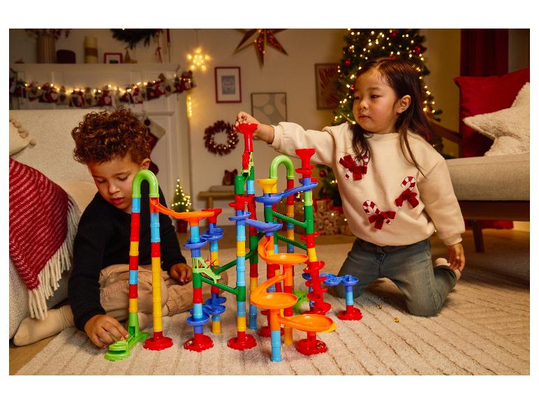 Dvě děti si hrají s barevnou kuličkovou dráhou na koberci ve vánočně vyzdobeném pokoji.