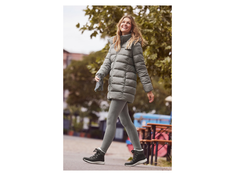 Žena v šedé zimní bundě, legínách a černých botách jde po ulici.