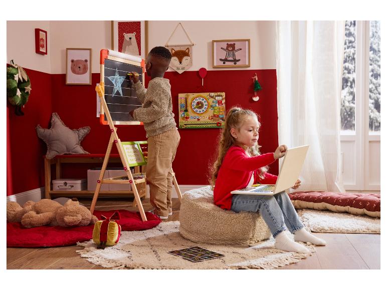 Dvě děti si hrají ve vánočně vyzdobeném pokoji: chlapec kreslí na tabuli, dívka používá notebook.