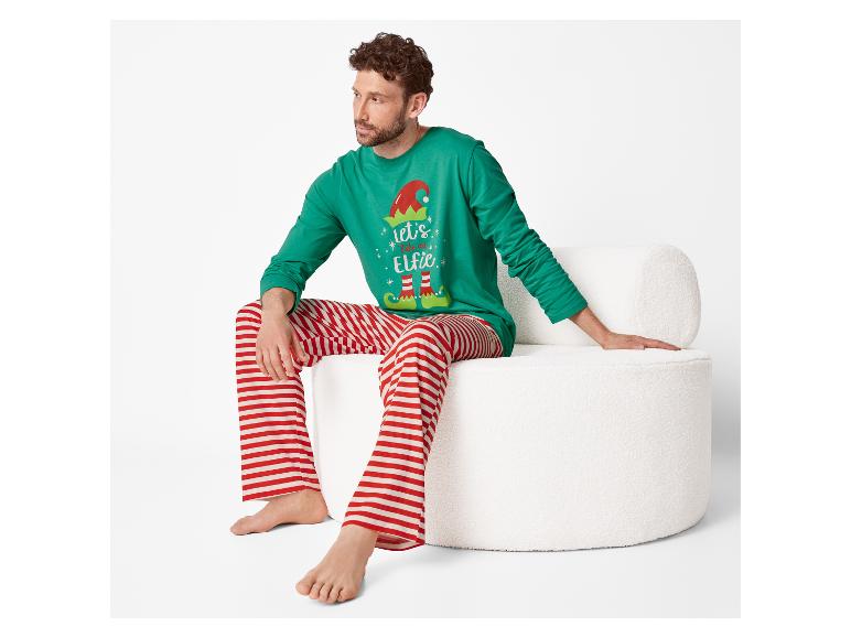 Muž sedí na bílé židli v zeleném vánočním pyžamu s nápisem „let's take an Elfie“ a červeno-bílými pruhovanými kalhotami.