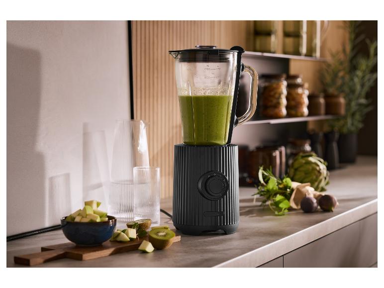 Černý mixér se zeleným smoothie, ovocem a bylinkami v kuchyni.