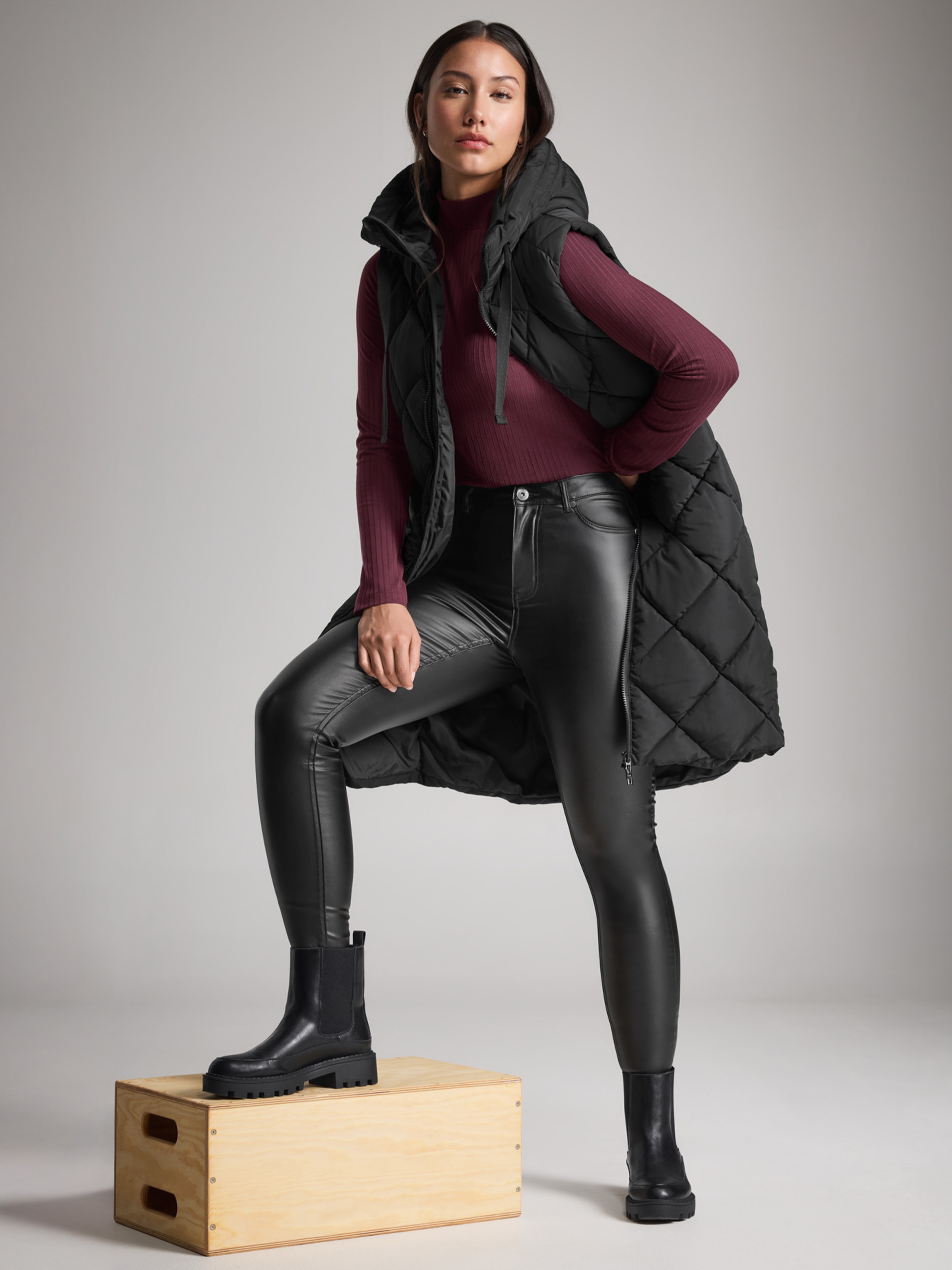 Žena v černé prošívané vestě, kožených džínách a kozačkách, s jednou nohou na krabici.