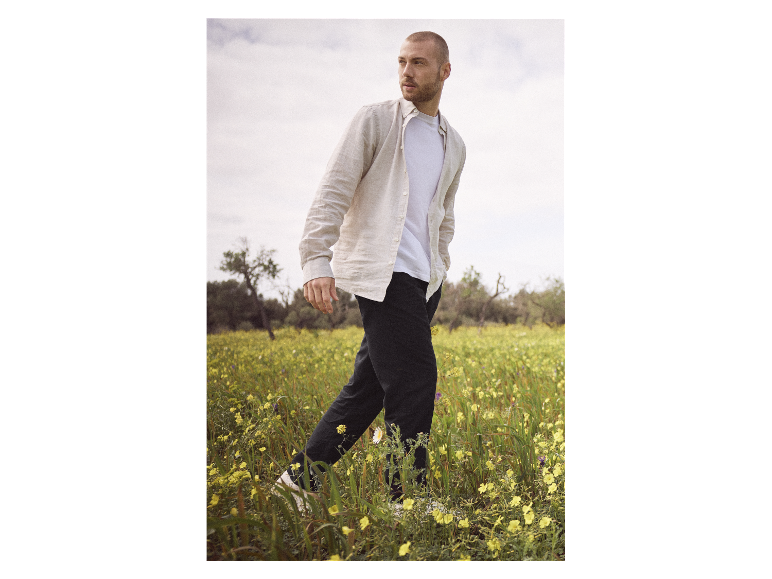 Muž jdoucí po poli květin, oblečený v lněné košili a kalhotách.