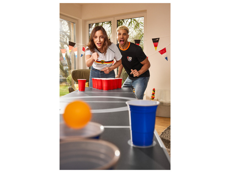 Dvě osoby hrají beer pong s oranžovým míčem a červenými kalíšky.