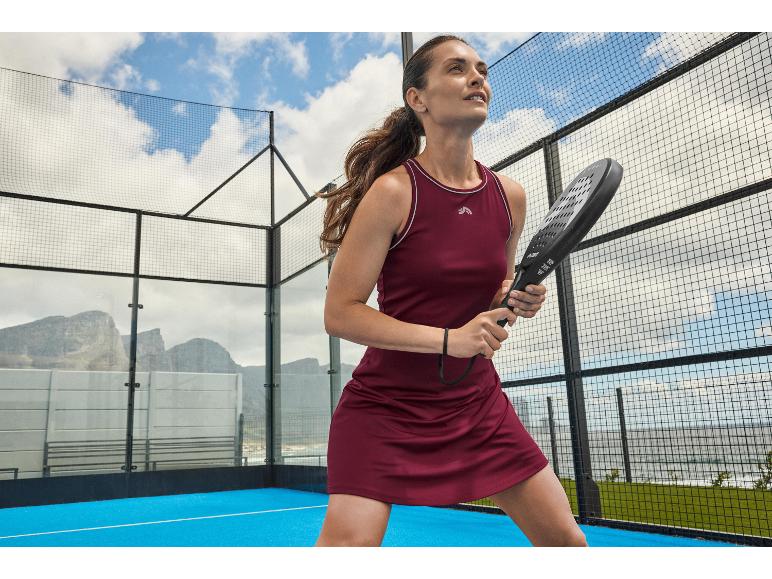 Žena v kaštanových padel šatech s raketou na padel kurtu s horami v pozadí.