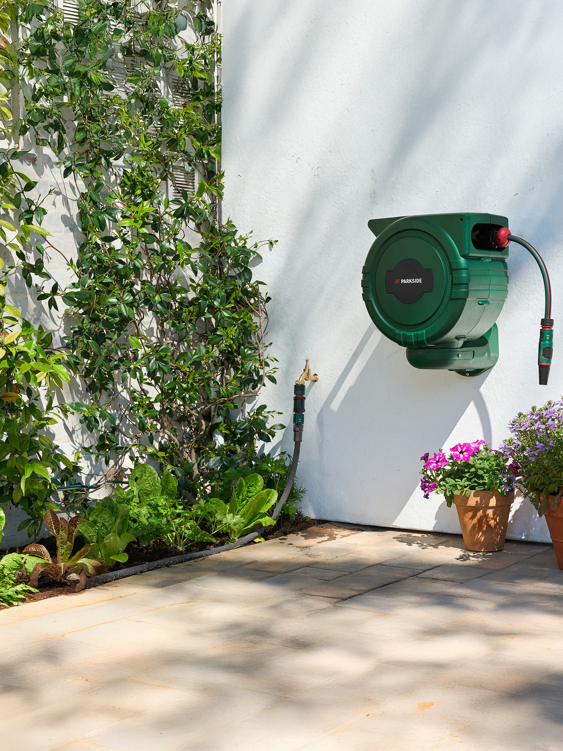 Zelený naviják na zahradní hadici PARKSIDE na bílé zdi s popínavými rostlinami a zeleninovou zahrádkou.