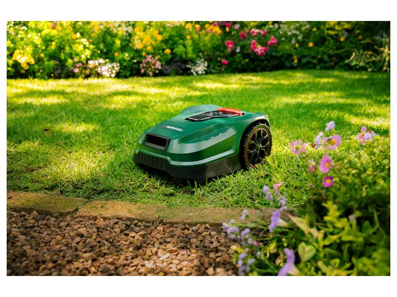 Zelená robotická sekačka na trávníku s květinami a keři v pozadí.