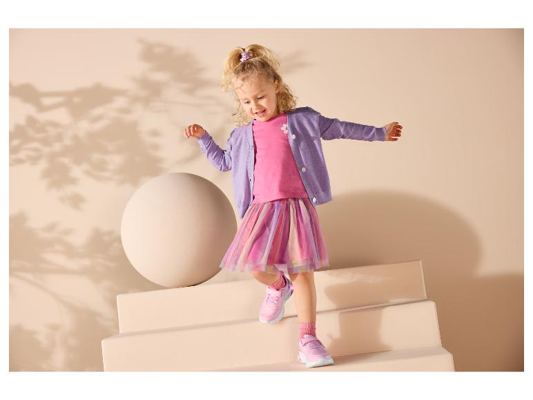 Usmívající se dívka v růžovém topu, fialovém kardiganu a barevné sukni, na sobě má růžové tenisky.