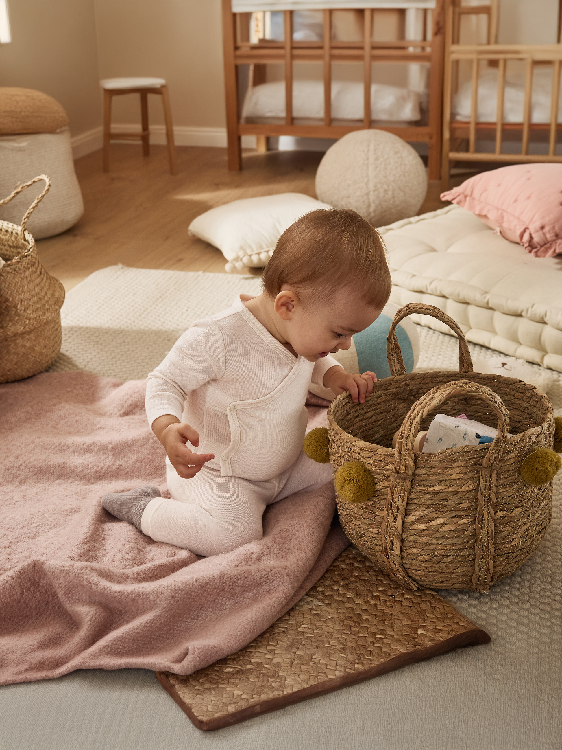 Miminko v bílém overalu si hraje s proutěným košíkem na růžové dece.