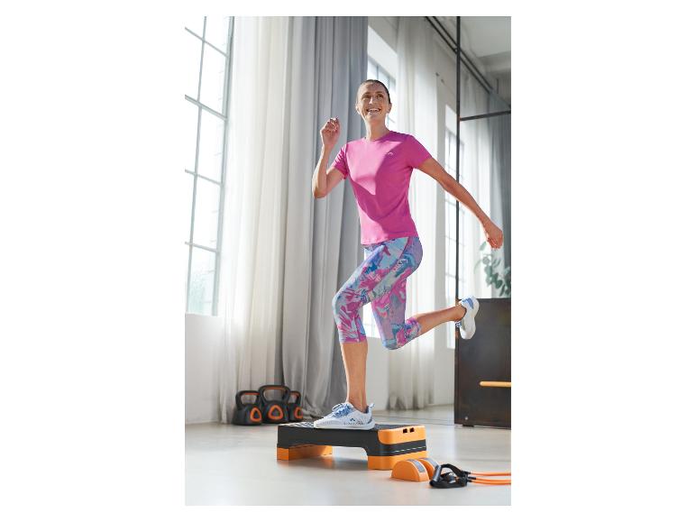 Žena v růžovém sportovním tričku a černo-oranžové step lavici cvičí