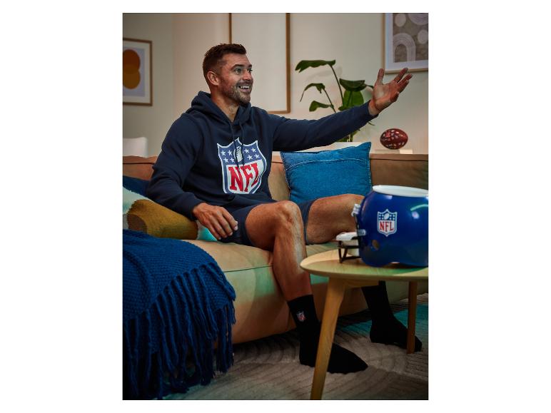 Muž v mikině a ponožkách s logem NFL sedí na gauči.
