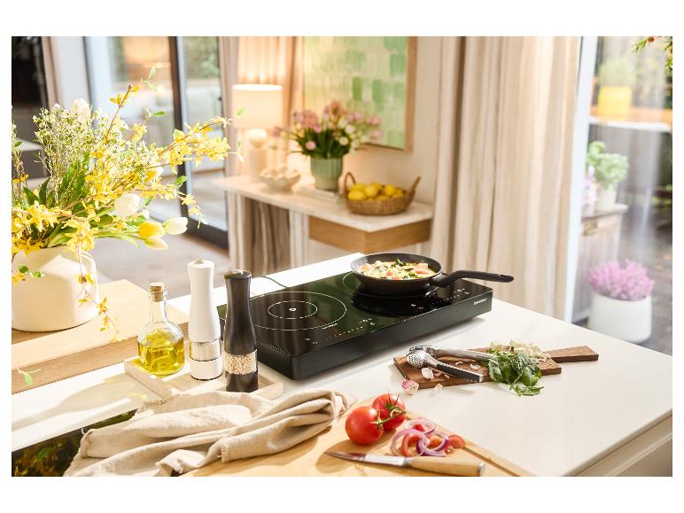 Černá indukční varná deska s pánví vařící zeleninu, obklopená čerstvými ingrediencemi.