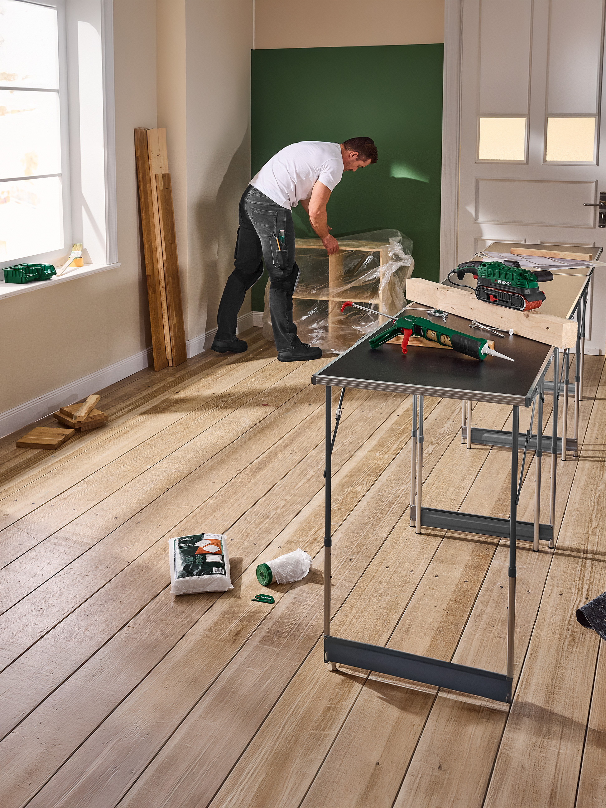 Muž pracuje na DIY projektu s nářadím Parkside, včetně brusky a pistole na tmel, na skládacích stolech.
