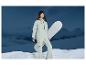 Žena v zeleném lyžařském overalu drží snowboard před zasněženými horami.