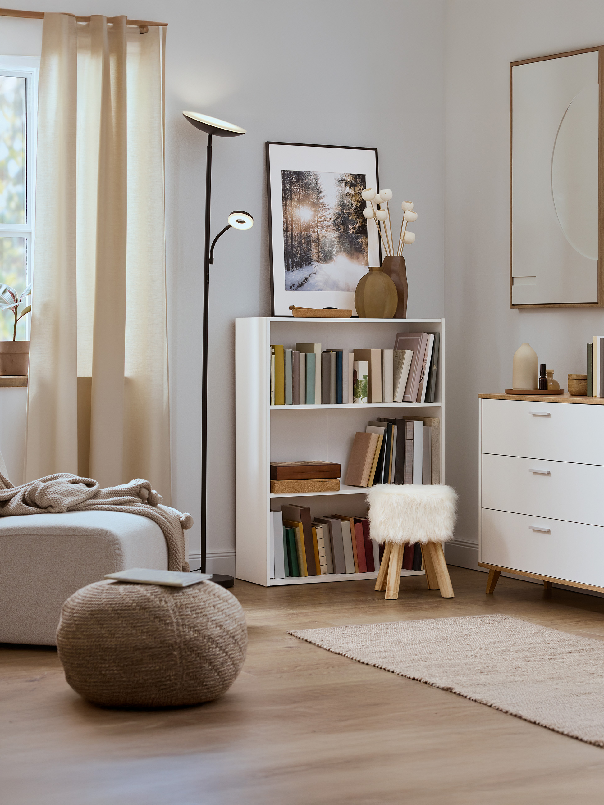 Světlý obývací pokoj s bílou knihovnou, pleteným pufem a moderní stojací lampou.