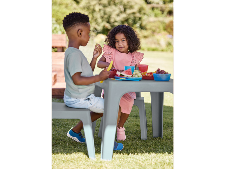 Dvě děti sedí u šedého dětského stolu a židlí venku na trávě.