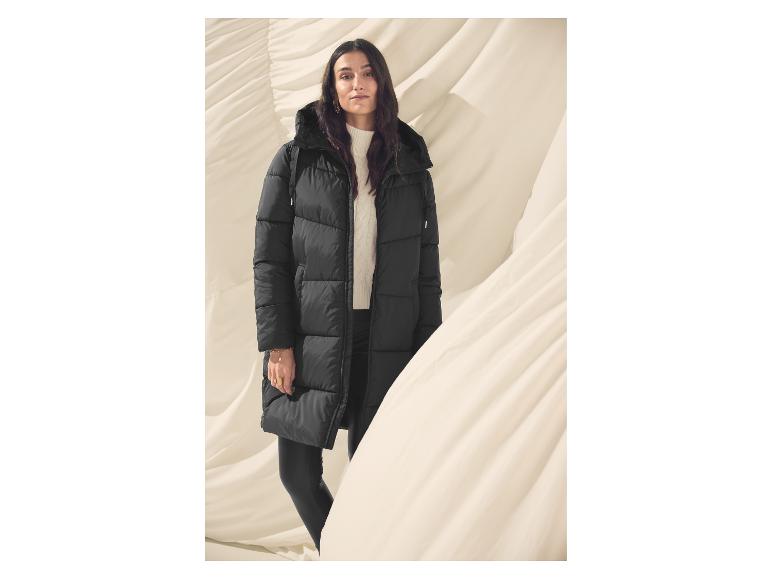 Žena v černé prošívané bundě a bílém svetru stojí před pozadím z drapované látky.