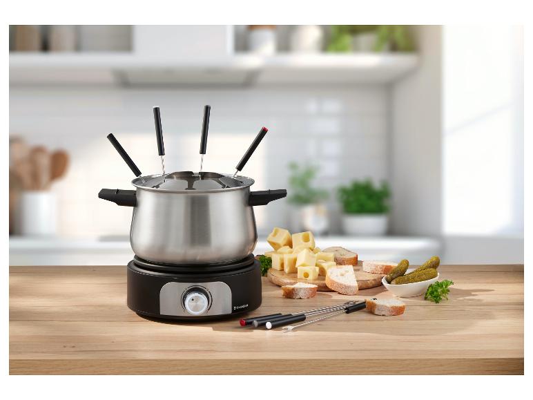 Fondue set se sýrem, chlebem a okurkami na dřevěném stole v kuchyni.
