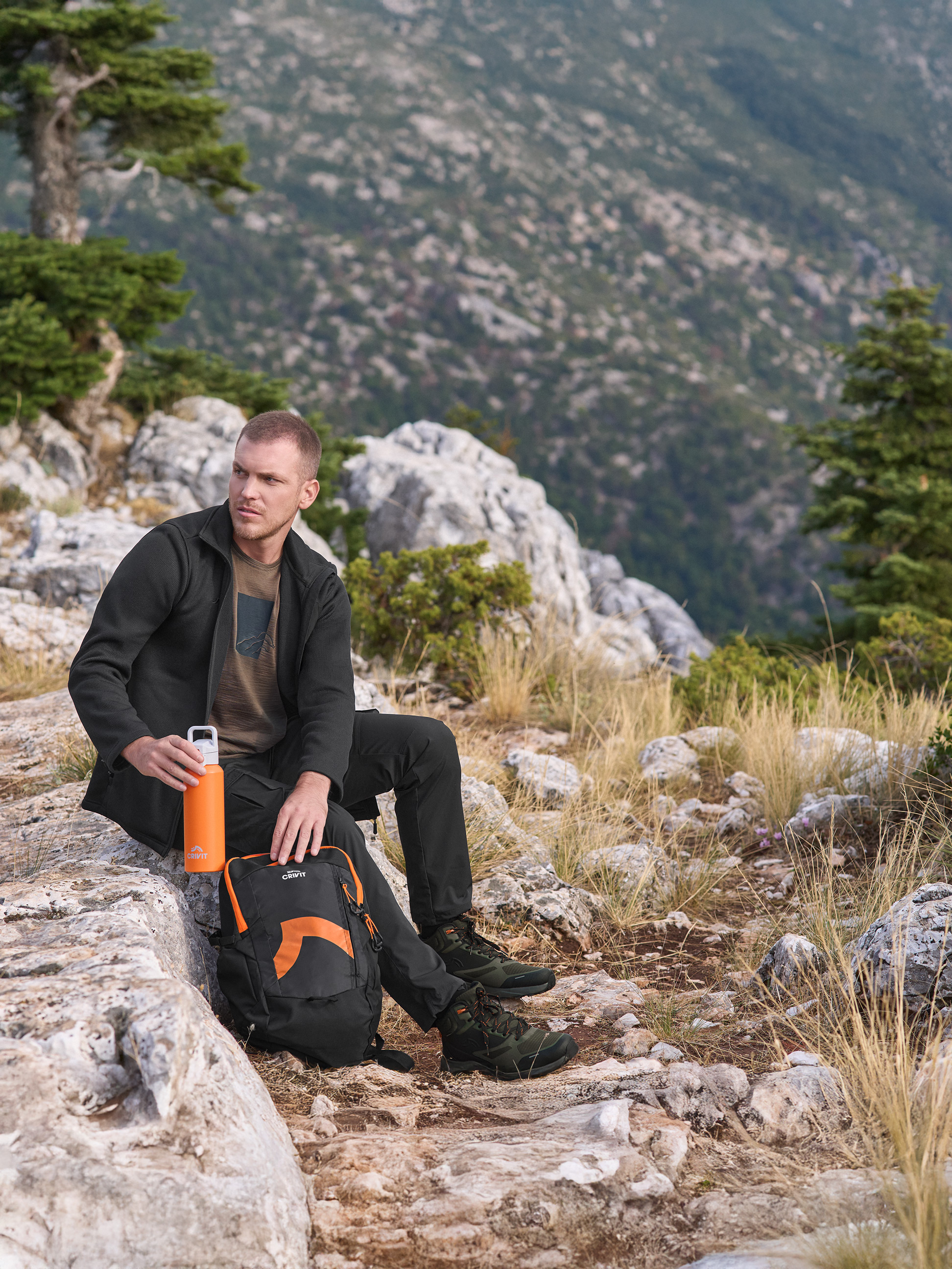 Muž v outdoorové bundě a kalhotách Crivit, drží oranžovou láhev na vodu Crivit, s batohem Crivit.