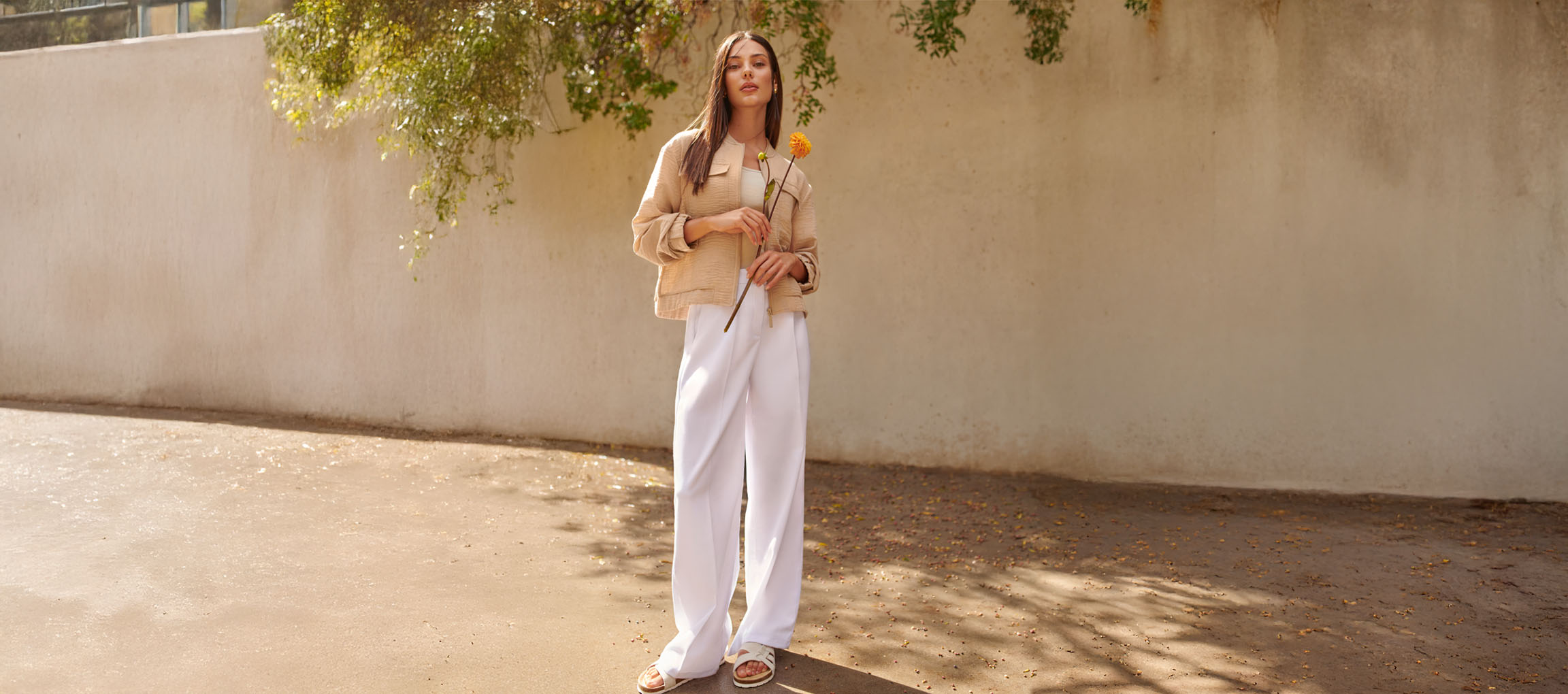 Žena v béžové bundě, bílých kalhotách a sandálech, držící žlutou květinu.