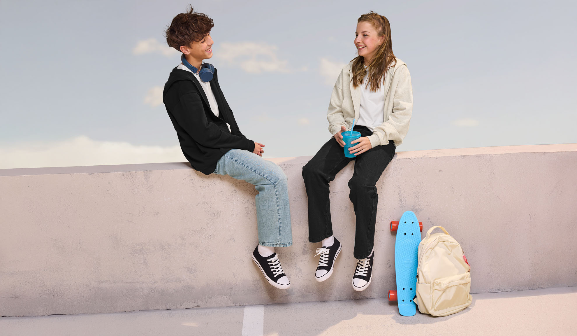 Dva teenageři v ležérním oblečení, sedící na zdi, s skateboardem a batohem poblíž.
