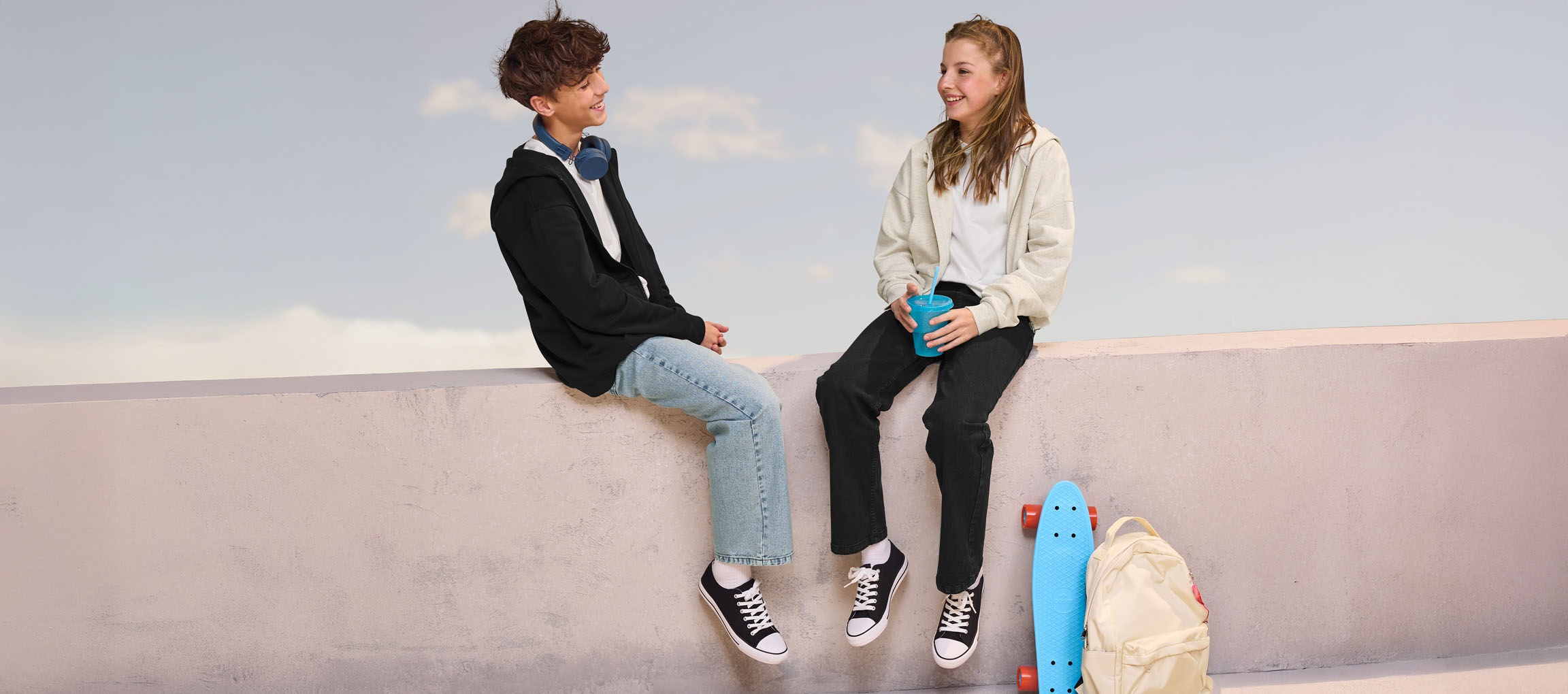 Dva teenageři v mikinách a džínách sedí na zdi, s skateboardem a batohem poblíž.