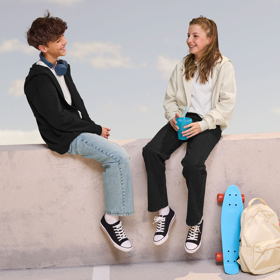 Dva teenageři v mikinách s kapucí a džínách sedí na zdi, vedle nich je skateboard a batoh.