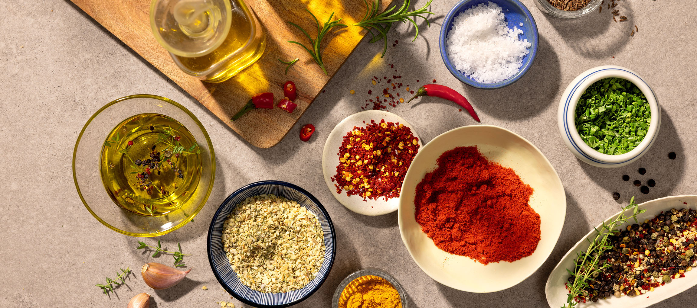 Různé koření a oleje, včetně papriky, chilli, soli, pepře a olivového oleje, na šedém pozadí.