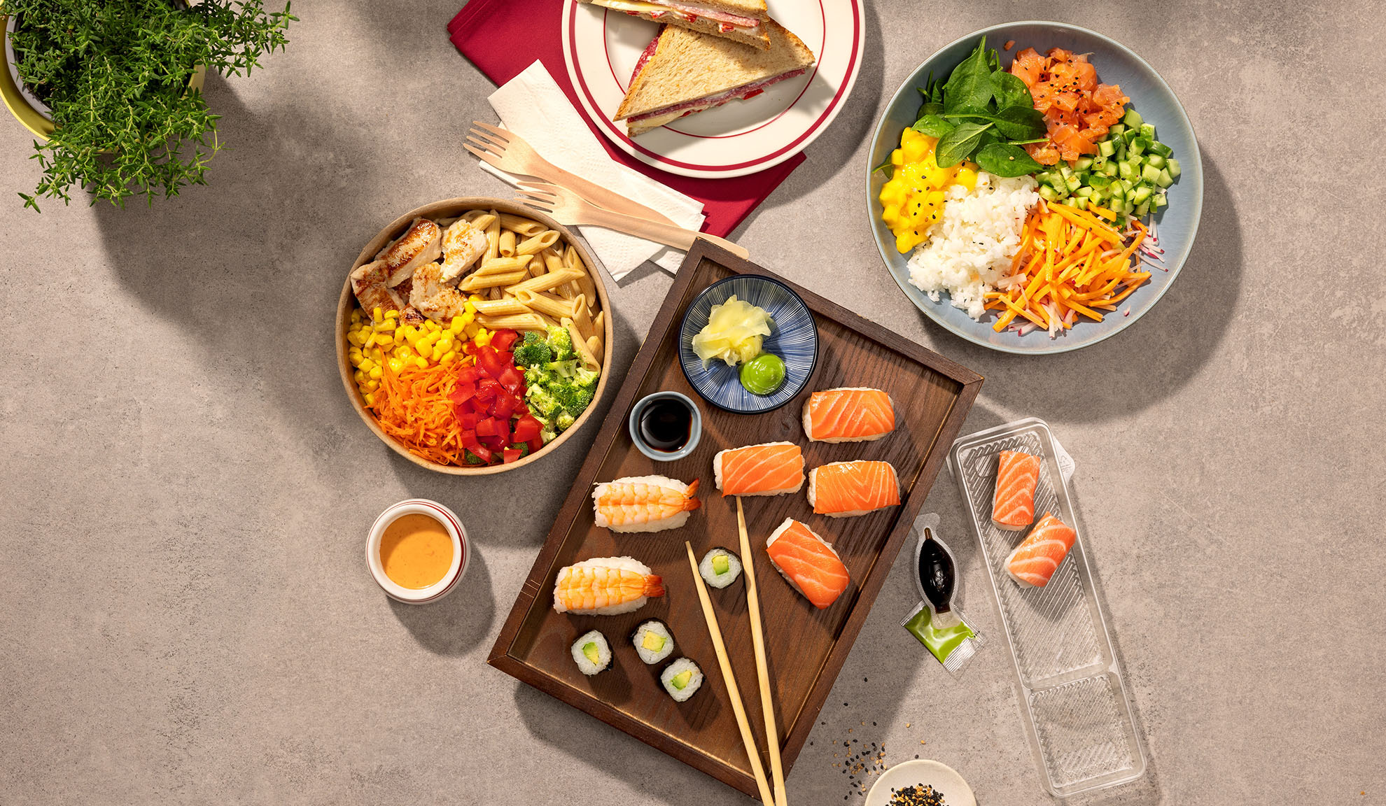 Pohled shora na různá jídla s sebou: sushi, poke bowl, těstovinový salát a sendviče.